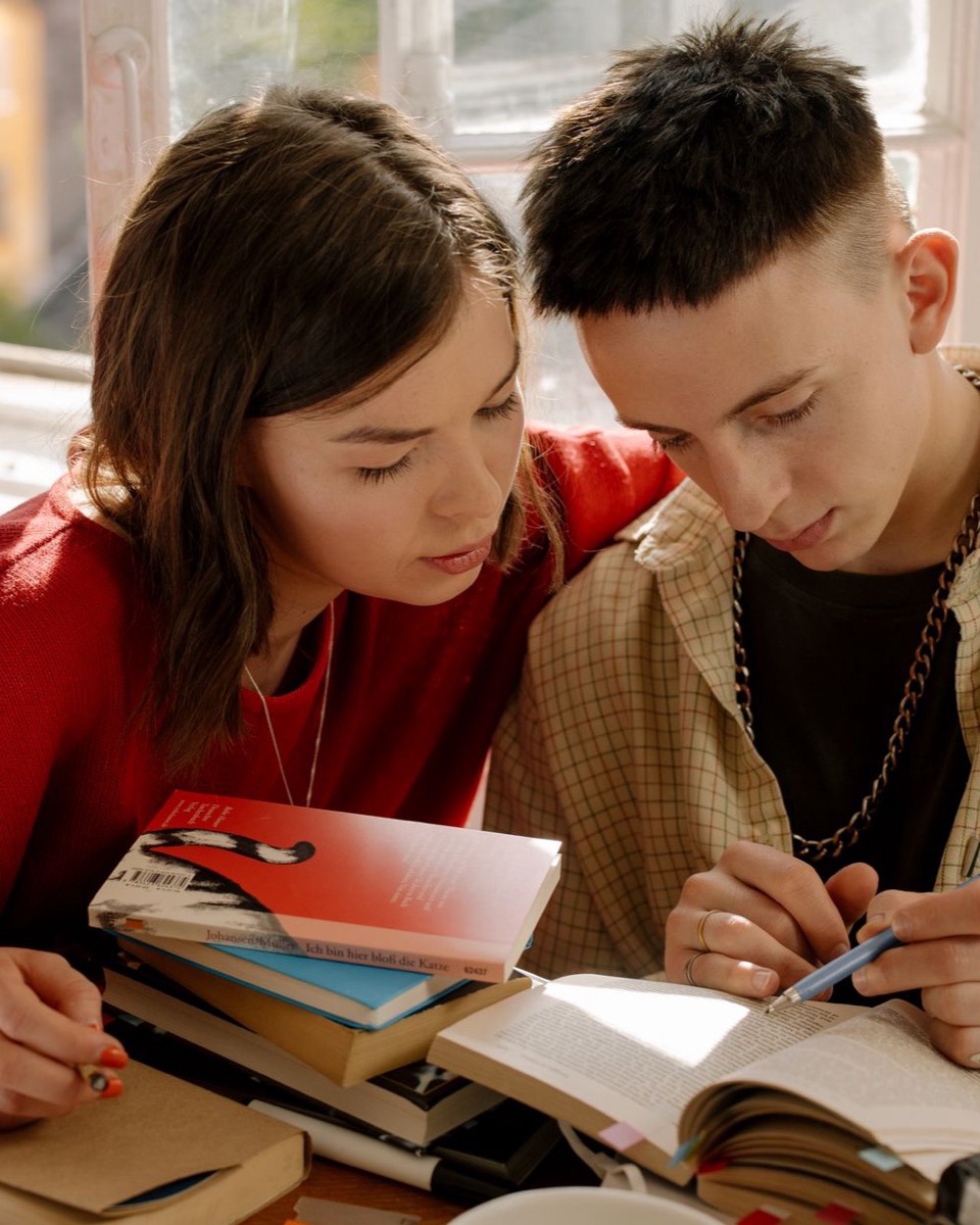Two students reading through a book together at a desk by a window